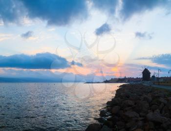Beautiful scenic sunrise on the coast of the Black Sea. The rays of the rising sun in the background of the sky with cumulus clouds. Silhouettes of houses and a mill. Old Nessebar in Bulgaria.