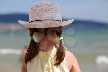 Little girl in white hat with tails looks at the sea on a bright sunny day. View from the back. Shallow depth of field. Focus on the model.