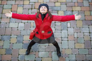 Happy young woman in a red coat arms outstretched and looking up against a background of paving slabs.
