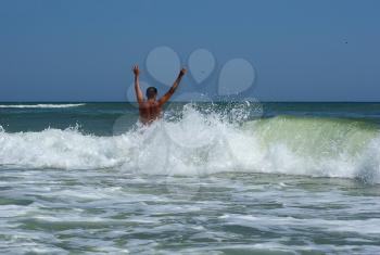 A man with his hands up his back to the camera on a background of a sea wave with spray foam. Young man bathing in the sea.