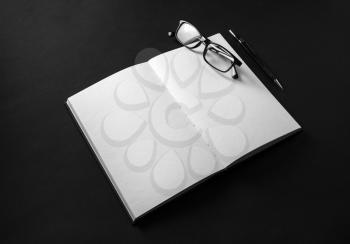 Mockup of opened blank book, pencil and glasses on black background.