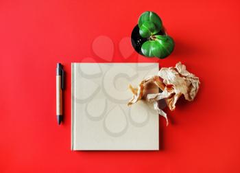 Closed book and blank stationery: pen, crumpled paper and plant on on red paper background. Top view. Flat lay.