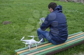 A man with a remote control quadroopter in his hands is sitting on a bench. White quadroopter prepare for flight