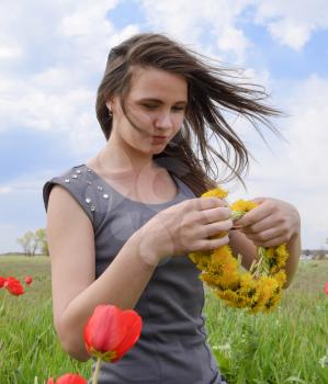 A girl with a wreath of dandelions in her hands. Beautiful fairy young girl in a field among the flowers of tulips. Portrait of a girl on a background of red flowers and a green field. Field of tulips