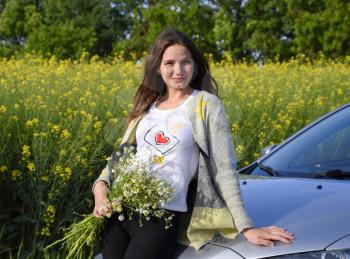 A beautiful young woman with a bouquet of daisies stands near a silver car.