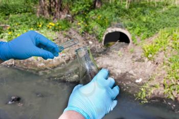 Check the water sample for infections. Hand in a glove with a bulb and a test tube against the background of a sewer pipe, dirty water. Pollution of ecology, environment