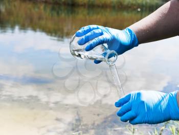 Fence, check the water sample for infections. Gloved hand with a flask and a test tube on a background of lake, river, sea. Pollution of ecology, environment
