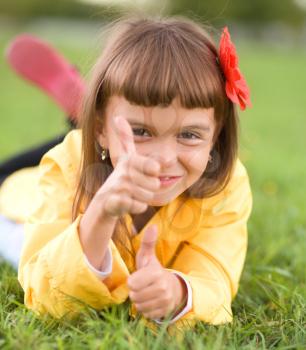 Little girl lying on green grass is showing thumb up gesture