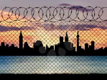 Concept of a border zone. Silhouette of the city behind a fence with barbed wire on a background of sunset and sea