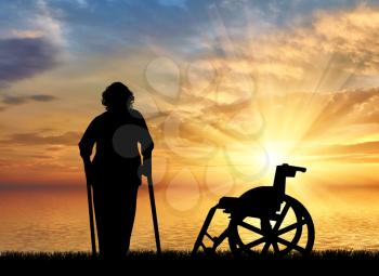 Silhouette of an old woman on crutches on a background of sea sunset and wheelchair. Concept of disability and old age