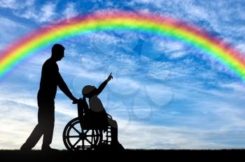 Silhouette of a disabled child girl sitting in a wheelchair and her dad showing a finger at a rainbow in the sky. Conceptual image of the life of children with disabilities