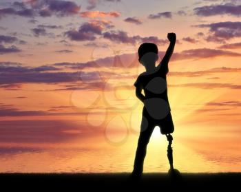 Children with disabilities concept. Happy disabled boy with a prosthetic leg standing at the sea at sunset