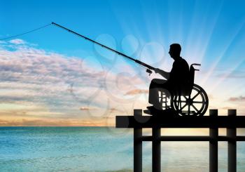 Silhouette of a disabled man in a wheelchair with a fishing rod in his hand fishing near the water on the pier. Concept of leisure and leisure of people with disabilities