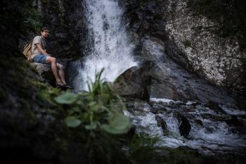 Young barefooted tourist wearing jeans short and backpack sitting on big rock looking peaceful and relaxed with waterfall on background