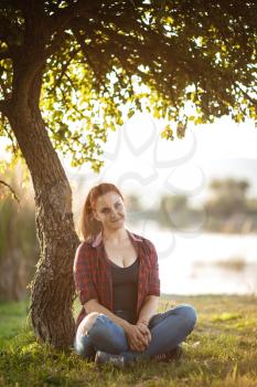 Attractive young woman enjoying her time outside in park with sunset in background, dressed in plaid shirt and jeans