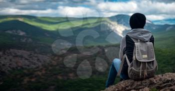 Young traveling woman wearing hat and sitting on the top of the mountain cliff with relaxing mood and watching beautiful view of woods and blue sky and clouds on vacation. Asian woman travel alone.