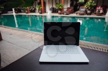 White laptop on the background of the pool in an exotic hotel. Conceptual workspace. Remote work, freelancer, internet, travel and vacation concepts.