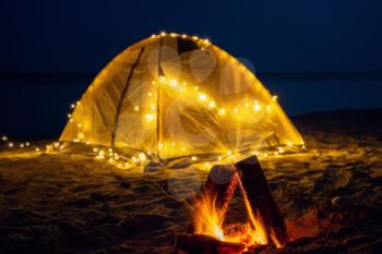 The fire at night on the beach. Summer mood. Night camping with a retro garland on sea shore.