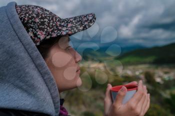tourist woman in a cap holds a mug with a hot drink in her hands, enjoying the scenery. Beautiful green valley and dramatic rain sky