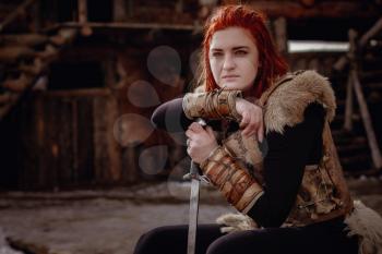 Viking woman with red hair. In the hands of the sword to fight the enemy. Reconstruction of a medieval scene. Against the backdrop of a large viking village.