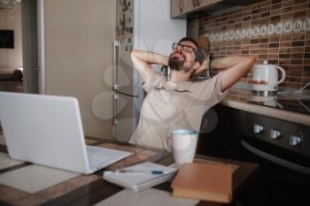 Smiling hipster freelancer holding hands behind head sitting at office desk behind laptop. Happy employee feeling no stress, relaxing, watching funny video after successful working