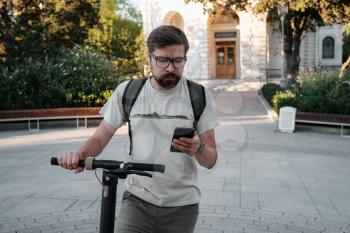 hipster man commuter with electric scooter outdoors in city, using smartphone. Young millennial guy using cellphone rental of eco transportation in summer