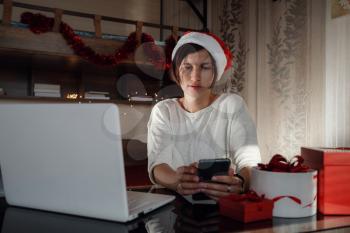 Concept of Christmas online shopping, buying gifts, congratulations over Internet. Cute lady sits with laptop, looks at the screen. Nearby gift boxes. white and red