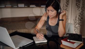 Young asian female student in headphones using laptop, writes notes . Cozy office workplace, remote work, E learning concept.