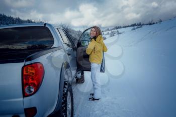 Young cheerful woman posing against the car and snow forest. Attractive woman dressed yellow jacket sitting at Passenger seat in silver car, winter season