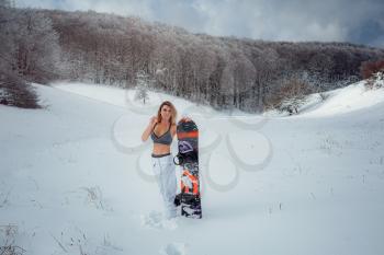 Female snowboarder hold snowboard and going to snowboarding. winter sport activity, forest snow outdoors lifestyle. Girl wearing a short top and ski white pants.