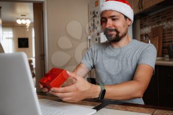 A hipster man with a red cup sitting on a sofa at home at Christmas time. Virtual Christmas house party. Online team meeting video conference calling from home. Man wearing Santa hat