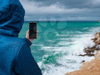 A woman take a photo of the sea on smartphone, female travels along the coast in the cold season