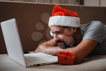 A hipster man with a red cup sitting on a sofa at home at Christmas time. Virtual Christmas house party. Online team meeting video conference calling from home.