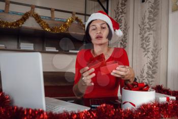 Excited asian Woman Holding And Opening Christmas Present, Smiling Lady Unwrapping Gift Box, Sitting On The table At Home In Living Room With Laptop Computer, Celebrating Online