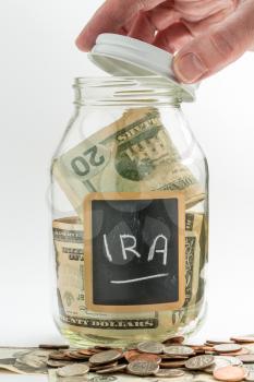 Hand opening lid of glass jar on white background with black chalk label and used for savings US dollar bills for IRA and retirement
