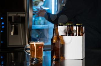 Six pack of brown beer bottles in plain white cardboard carrier with mug of ale on stainless steel kitchen or bar counter. Open fridge or refrigerator out of focus in rear.