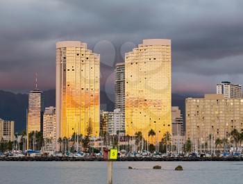 Tall apartment blocks in Honolulu reflect the setting sun in their mirrored windows as the sunset illuminates the clouds behind