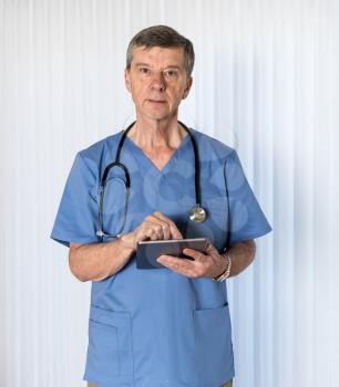 Senior male caucasian doctor with stethoscope in medical scrubs and checking notes on smartphone tablet device