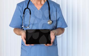 Senior male caucasian doctor with stethoscope in medical scrubs looking up and holding electronic tablet for message