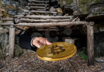 Miner engaged in mining for new bitcoins holds out a coin out of the entrance to an old coal mine