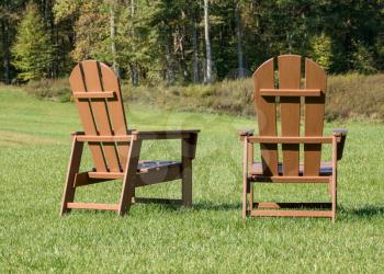 Rear view of the backs of a pair of adirondack wooden chairs facing a forest over grass lawn