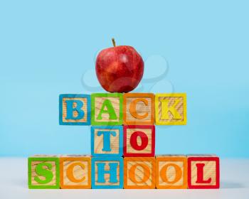 Stack of wooden blocks stacked to spell Back to School with red apple on top against blue background