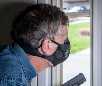 Senior man with face mask looking worried at front door and ready to defend home with a gun during coronavirus quarantine
