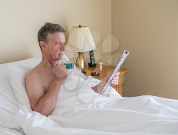Senior retired caucasian man lying in adjustable bed on incline. He is reading from magazine with cup of coffee
