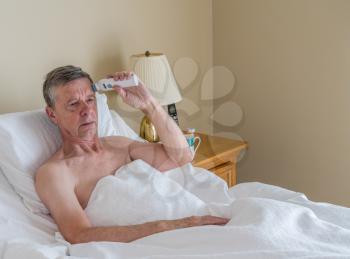 Senior retired caucasian man lying in adjustable bed on incline. He is checking his temperature for fever or signs of virus