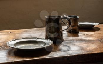 Antique pewter drinking mugs and flat plates with no food on large wooden dining table