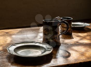 Antique pewter drinking mugs and flat plates with no food on large wooden dining table