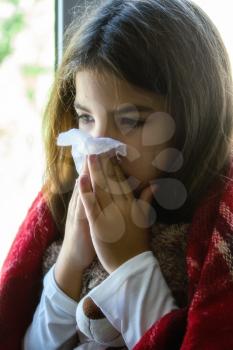 a sick little girl sits at the window wrapped in a blanket and blows her nose into a disposable napkin