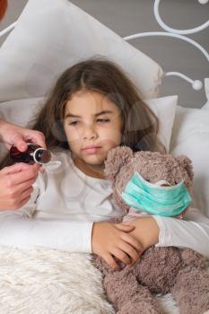 A little girl with a favorite teddy bear on whom they put on a gauze bandage is lying sick in bed and her mother is drinking her medicine.