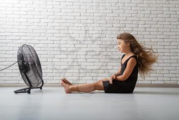 little girl struggling with heat and sitting in front of a fan with flying hair in an apartment on a bricky light wall background